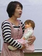 寸劇女性（妻）が赤ちゃん（人形）を抱っこしながら話しかけている様子