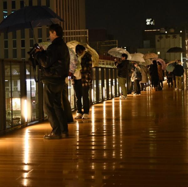 雨の降るオープンデッキに傘を差した複数の人が写っています。